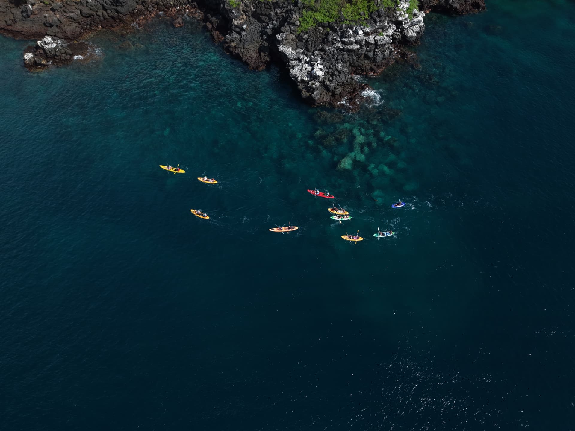 Aerial view of a group of kayakers spread across turquoise water below volcanic cliffs
