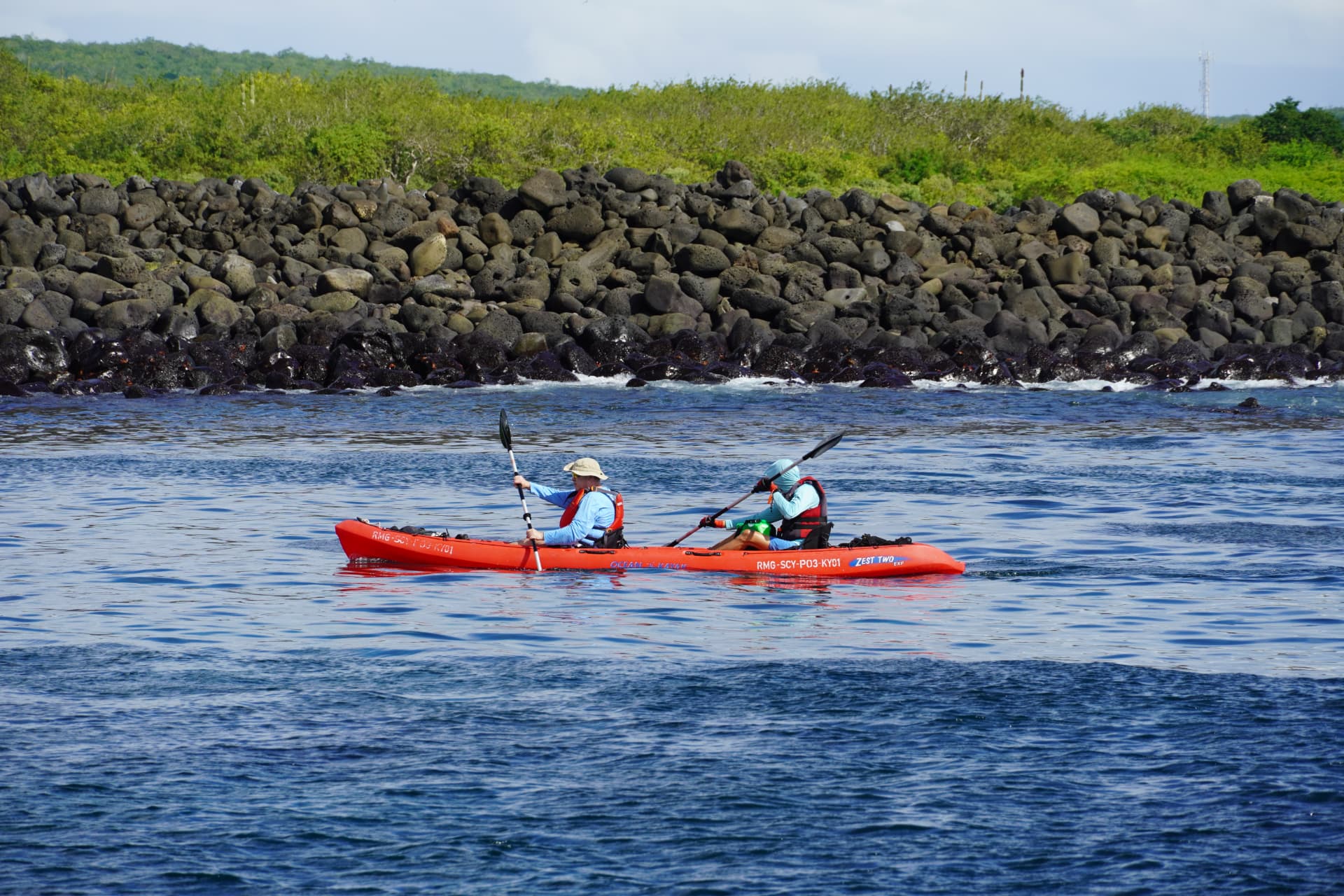 Two kayakers paddling a red double kayak along a black boulder shoreline